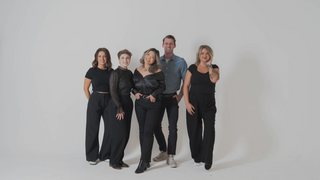 A group of five individuals posing together in stylish black outfits against a plain background.